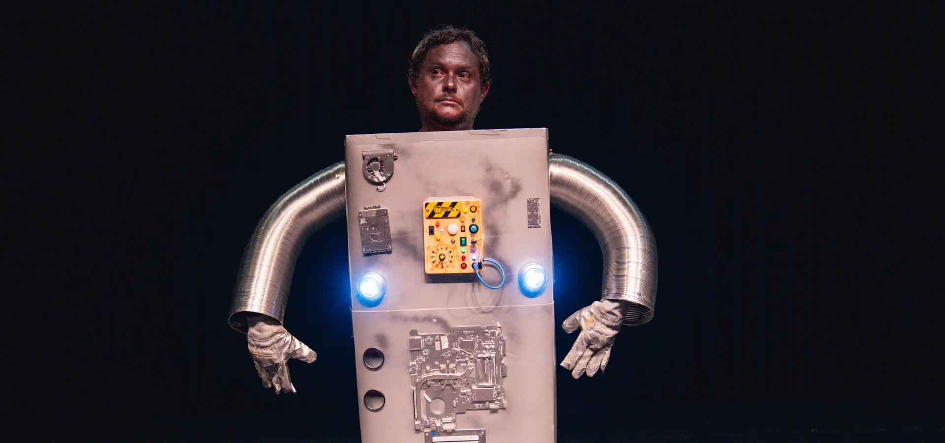 Persoon verkleed als robot met een kostuum van een grote rechthoekige doos met knoppen en tekeningen erop, op een donker toneel verlicht door theaterlicht.