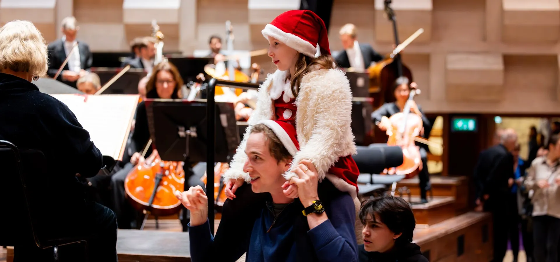 Een vader met twee kinderen in een concertzaal, waarvan één op zijn schouders zit met een kerstmuts op. Op de achtergrond repeteert een orkest, en de sfeer is feestelijk en warm.